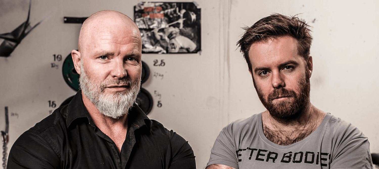 Two men are standing side by side indoors. One is bald with a beard, wearing a black shirt. The other has short hair and a beard, wearing a gray T-shirt.